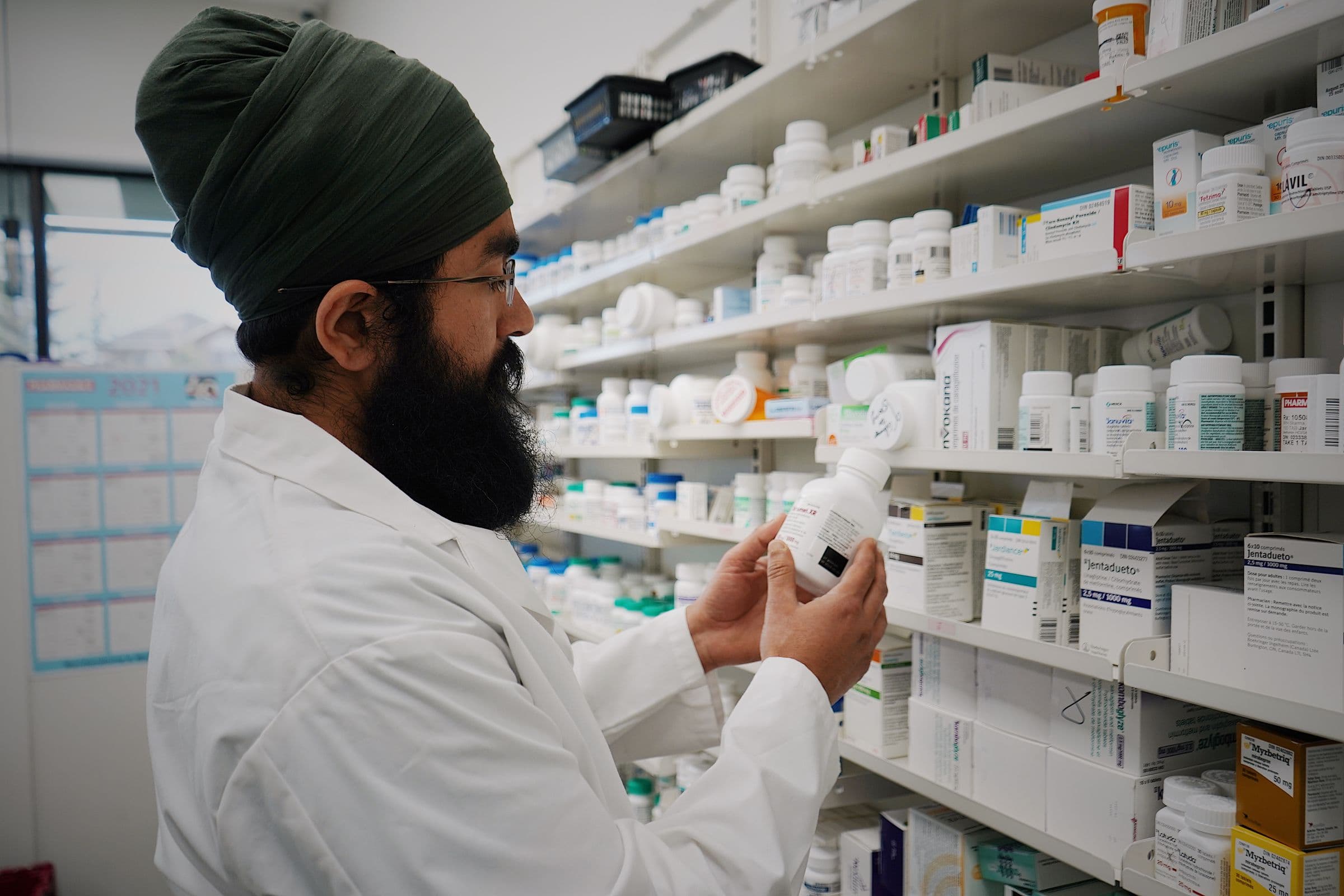 A pharmacist in a white coat checking a medication bottle against stocked clinical pharmacy shelves